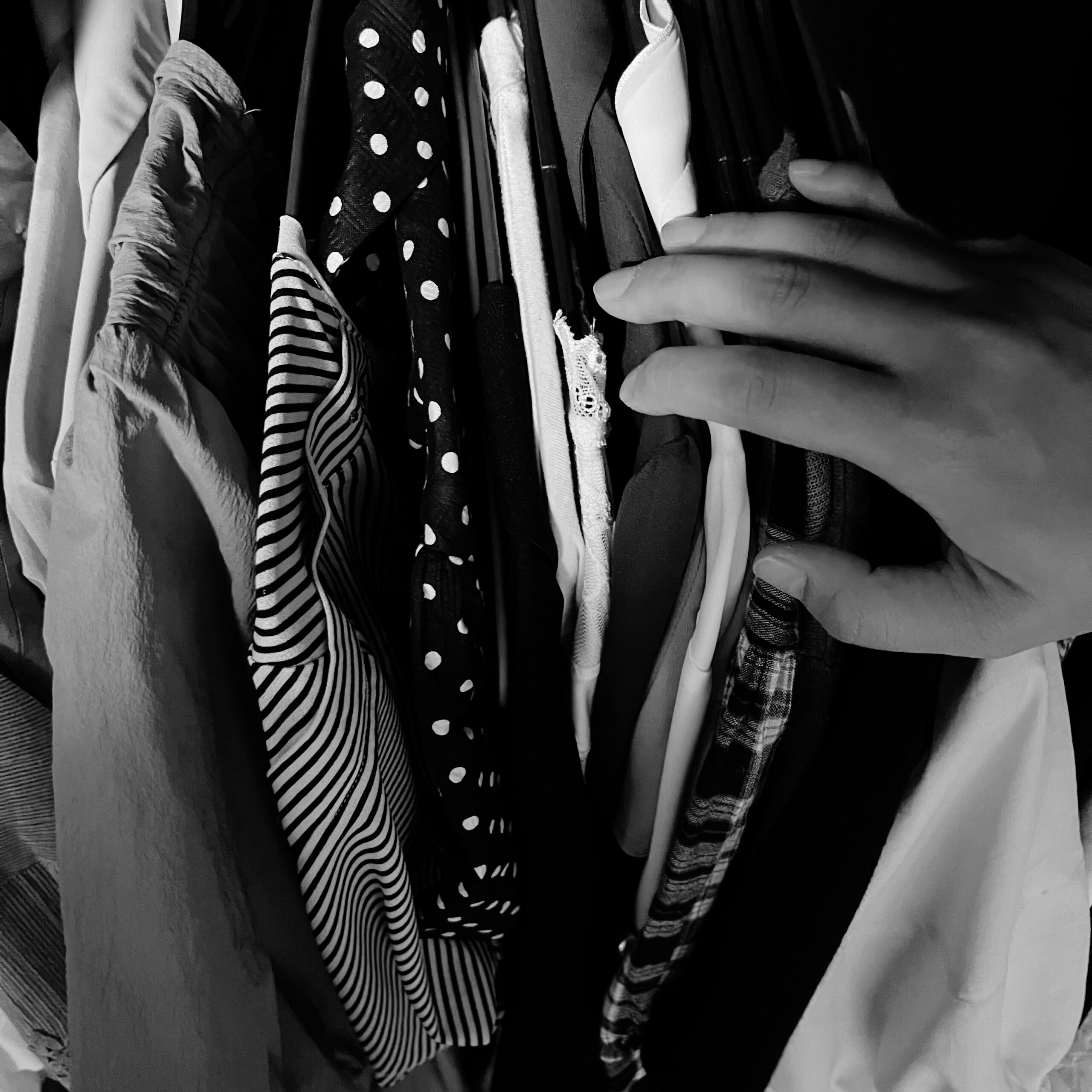 Mano eligiendo entre varias prendas colgadas, con distintos estampados y texturas, en una fotografía en blanco y negro.
